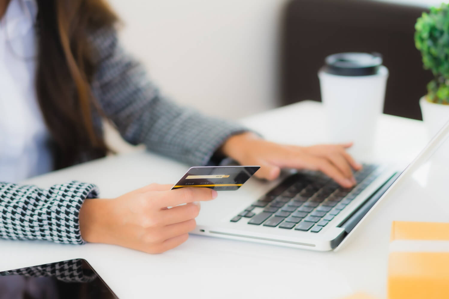 Person holding a credit card while using a laptop on a white desk with a coffee cup and plant nearby.