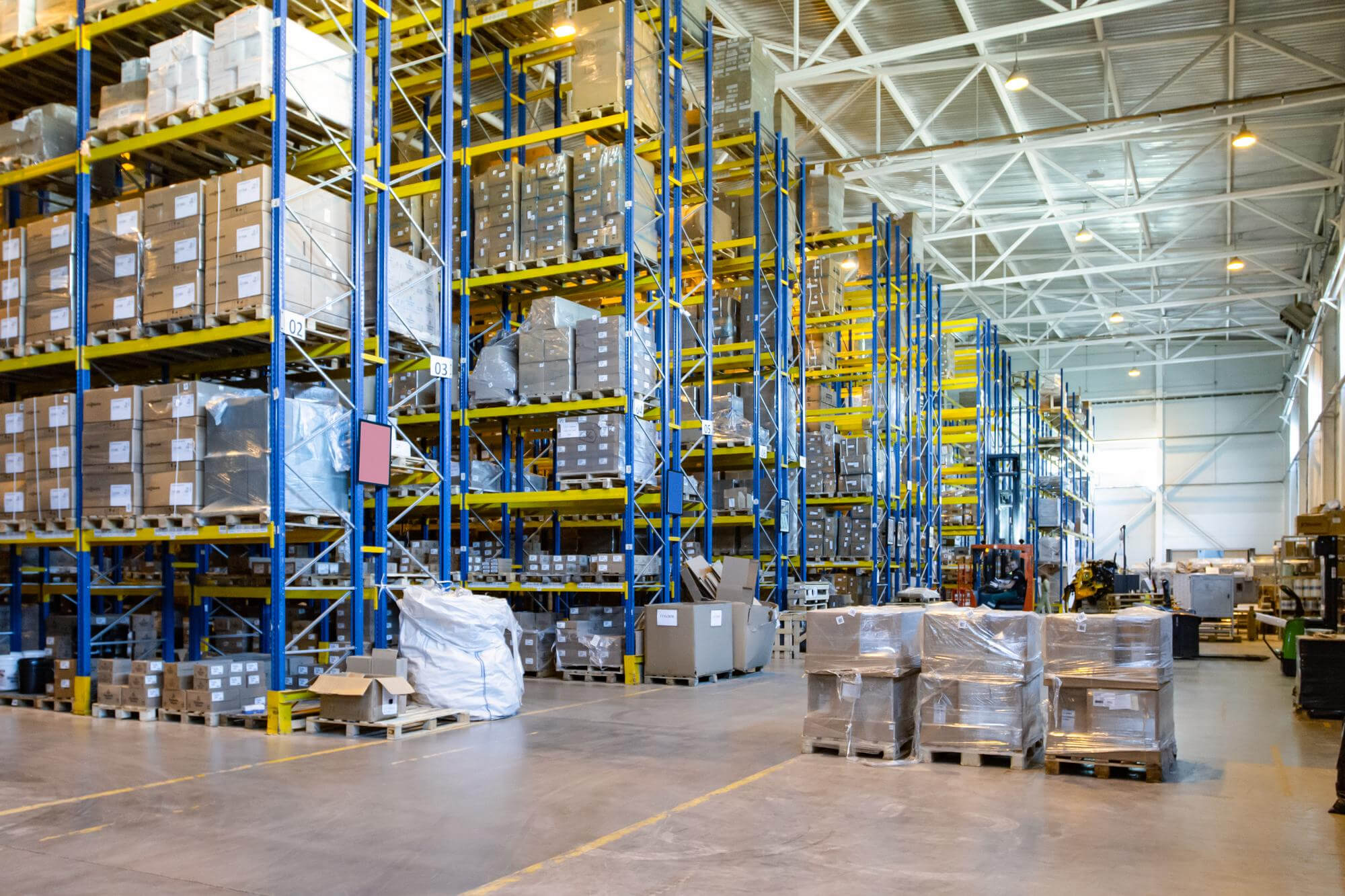Interior of a large warehouse with tall blue and yellow shelving units stacked with cardboard boxes on pallets and wrapped goods on the floor.