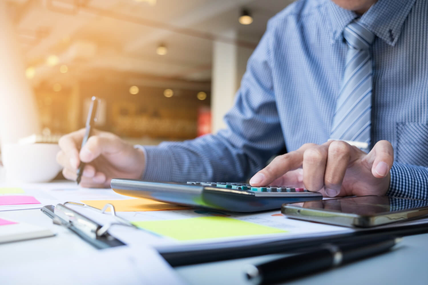 Man in a blue checkered shirt and striped tie using a calculator and writing with a pen at a desk with documents and a smartphone.