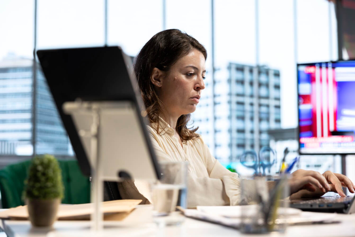 Woman focused on typing on a laptop at a desk in a modern office with large windows.