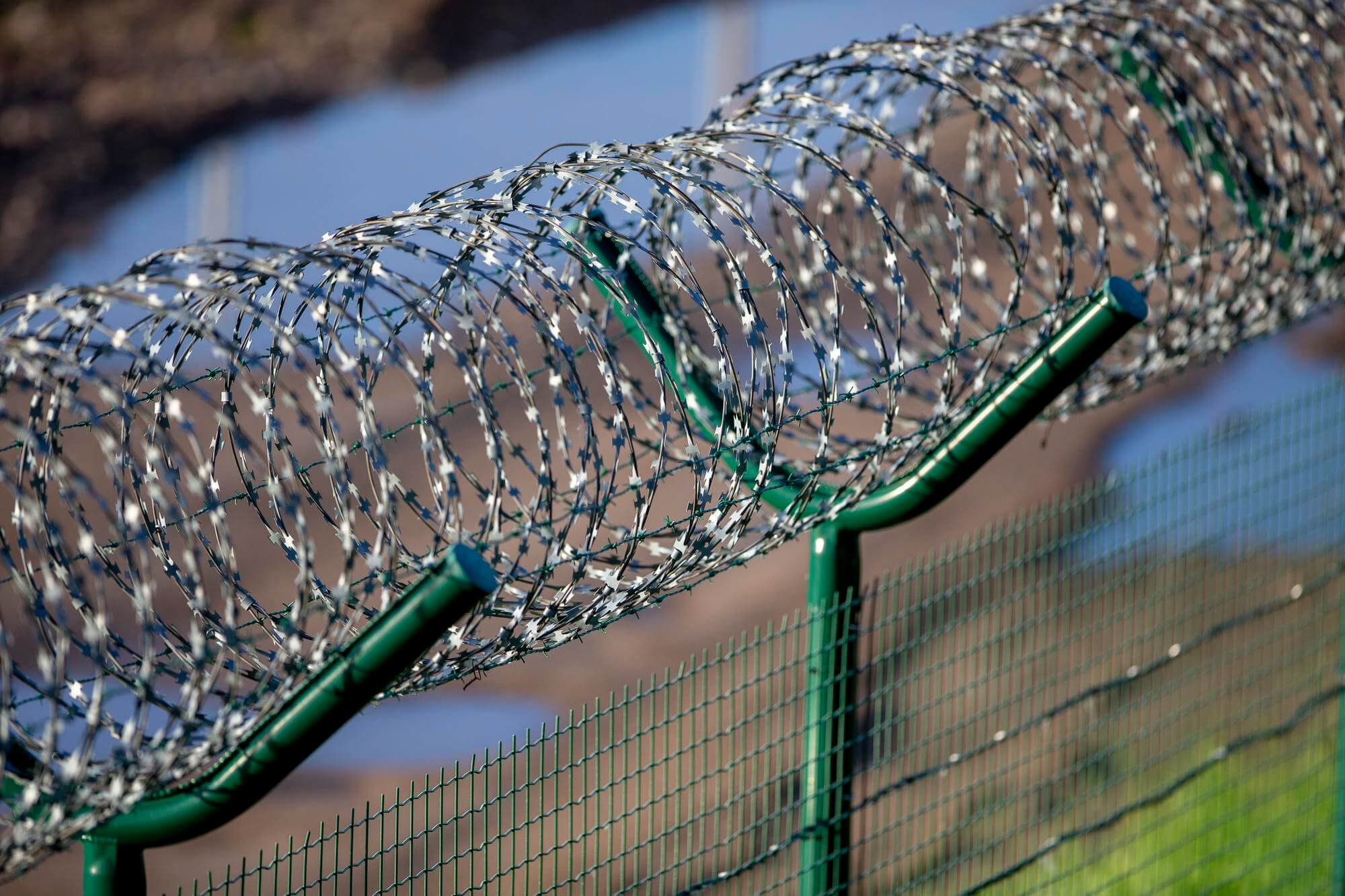 Close-up of coiled razor wire mounted on angled green metal posts above a green security fence.