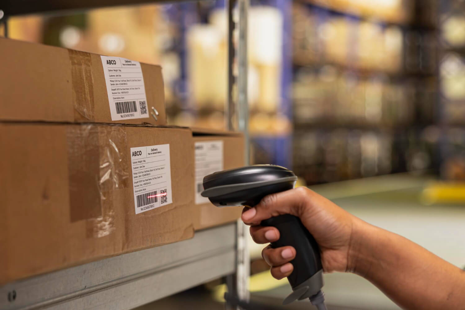 Hand holding a barcode scanner scanning labels on stacked cardboard boxes in a warehouse.