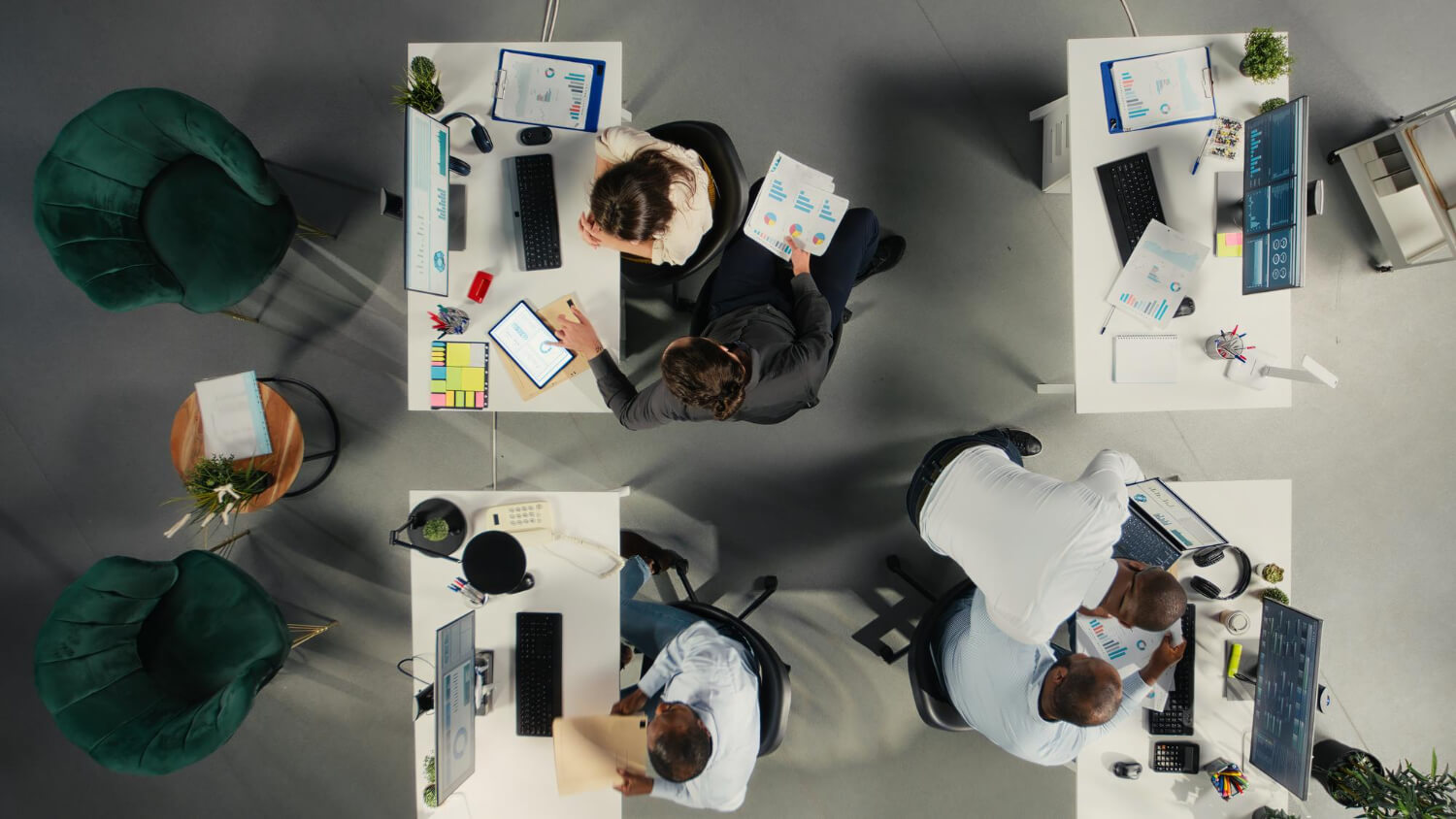 Overhead view of four office workers collaborating at adjacent white desks with computers, charts, and documents.