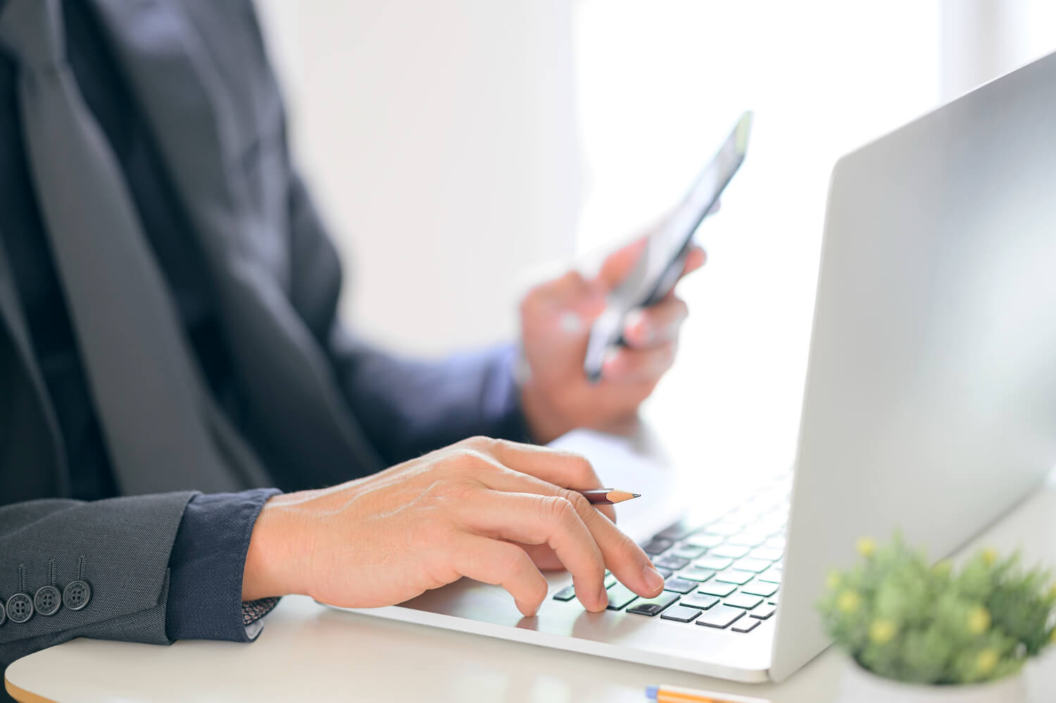 Person in business attire typing on a laptop with one hand while holding a smartphone in the other.