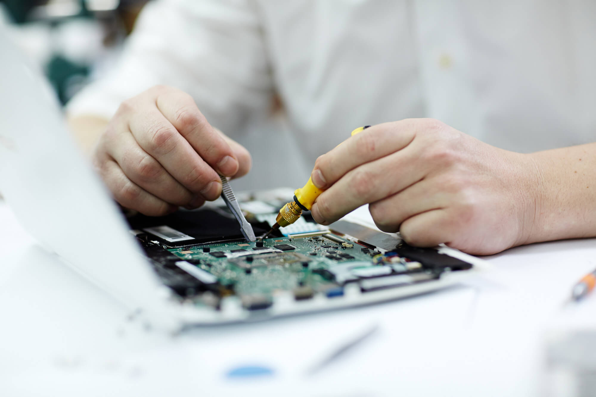 Close-up of hands repairing a laptop motherboard using tweezers and a small screwdriver.