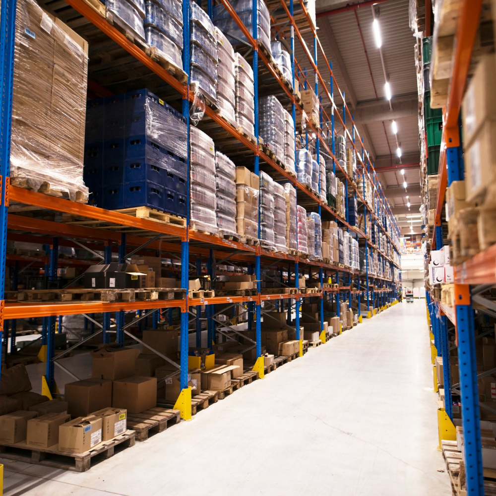 Long aisle of a large warehouse with tall shelves stocked with pallets of wrapped goods and cardboard boxes.