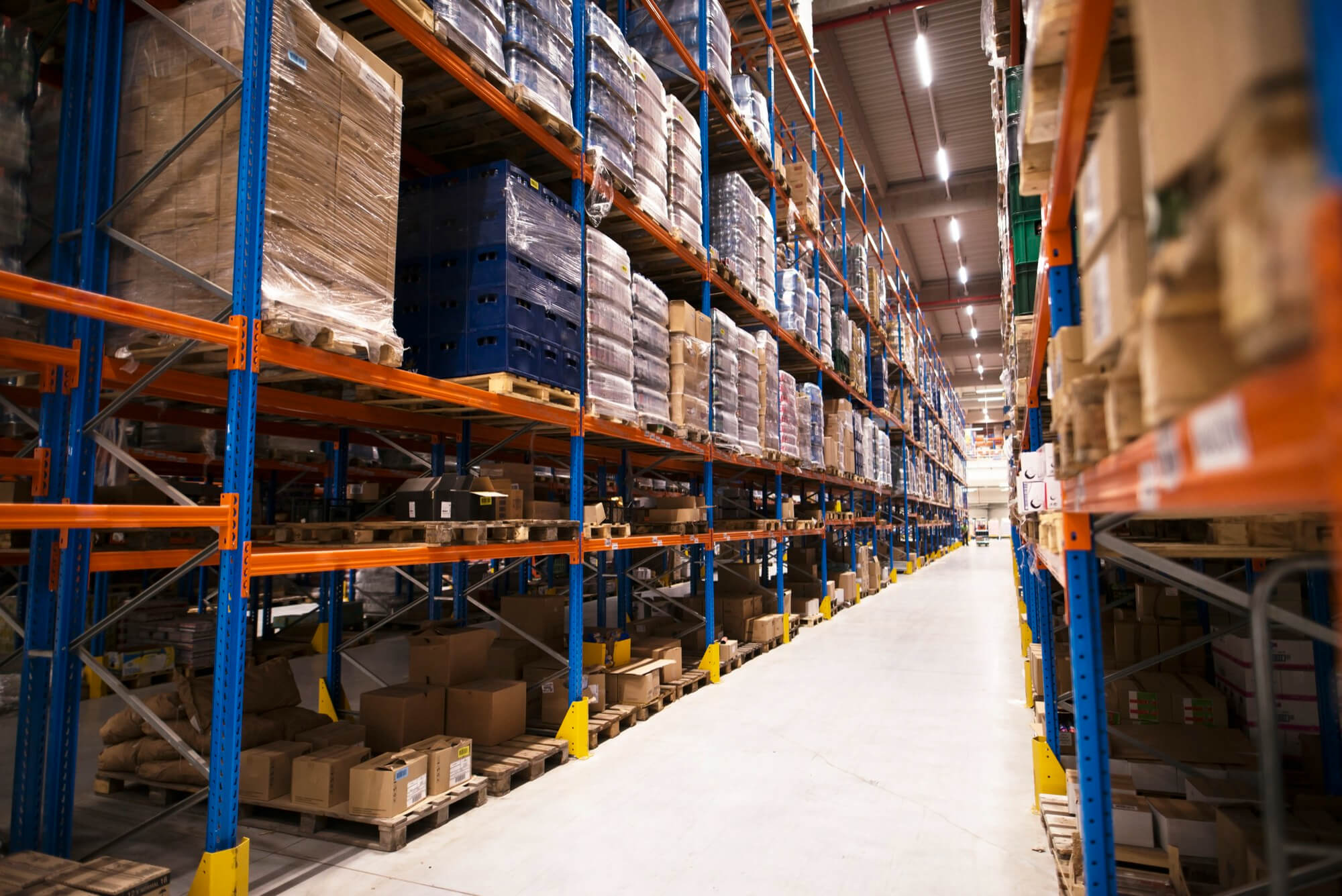 Wide aisle in a large warehouse with tall blue and orange metal shelves stocked with pallets of boxed goods.