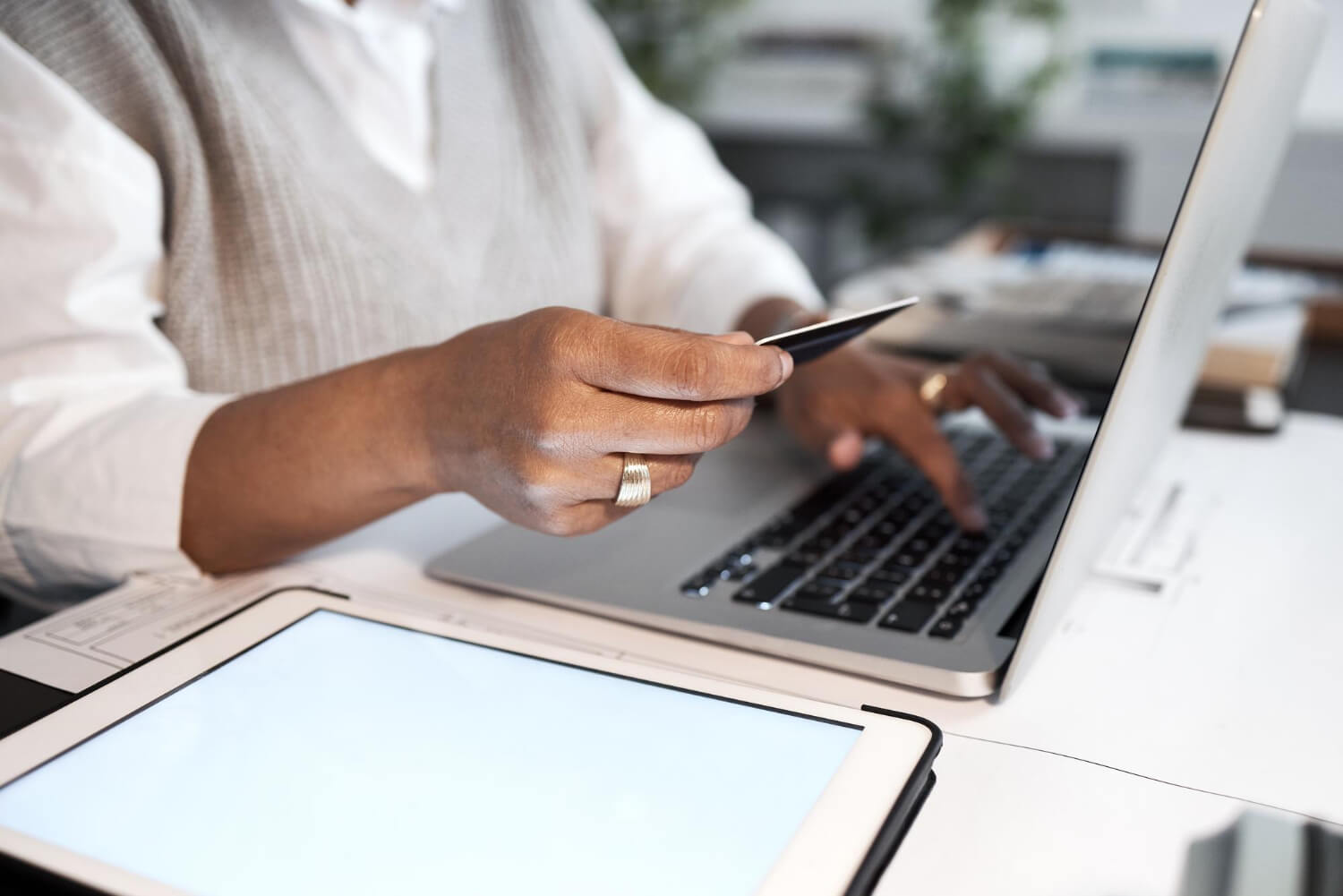 Person holding a credit card and typing on a laptop keyboard at a desk with a tablet nearby.