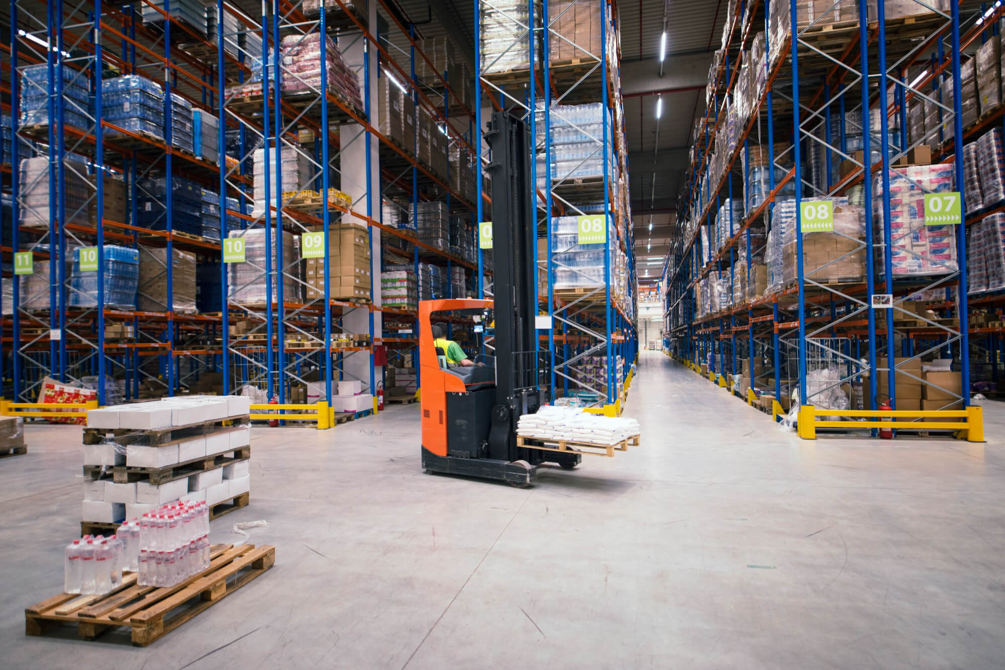 Forklift moving a pallet of goods inside a large warehouse with tall, fully stocked shelves.