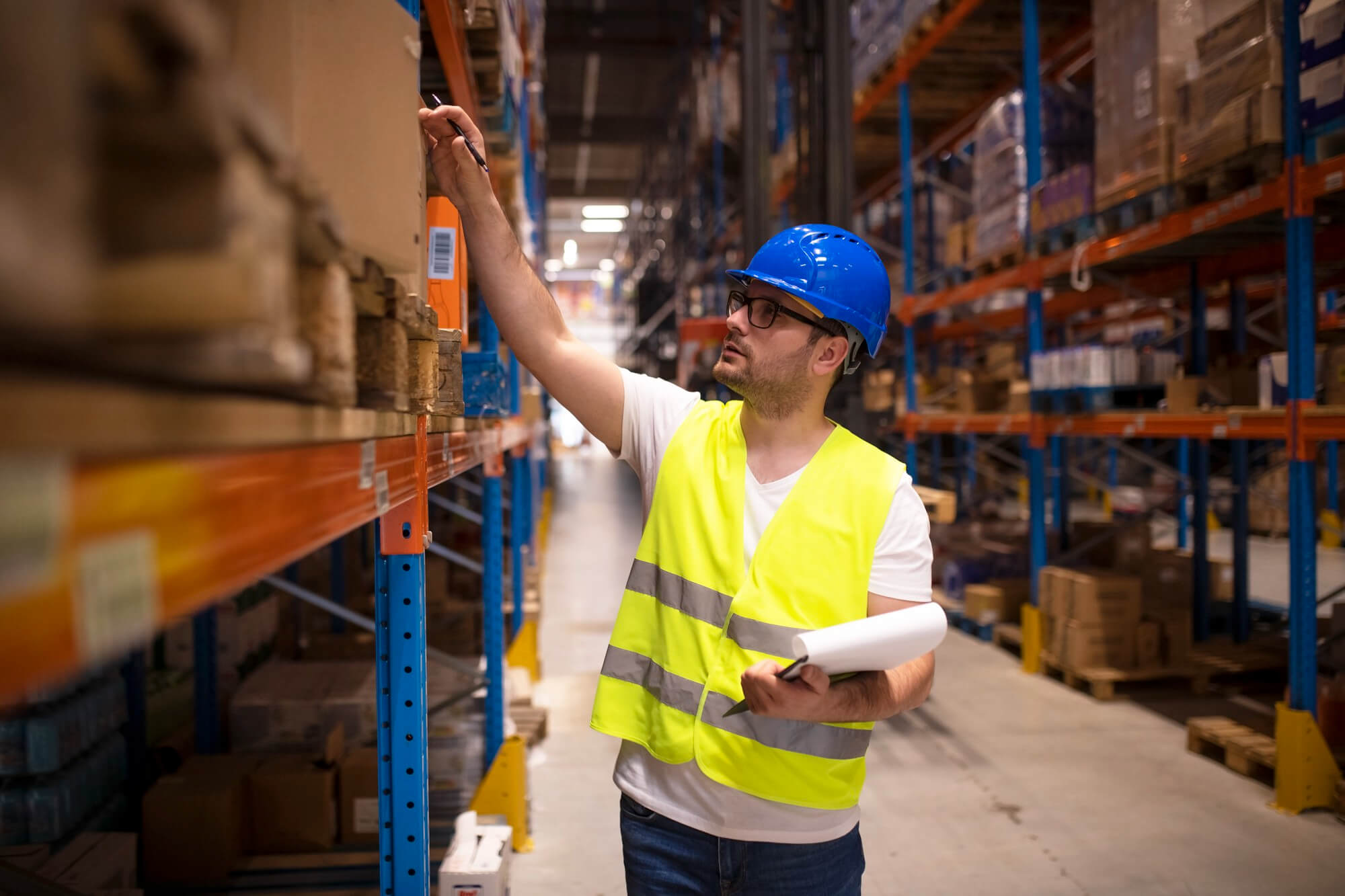 Warehouse worker in a blue hard hat and yellow safety vest checking and writing on a box in an aisle filled with shelves and pallets.