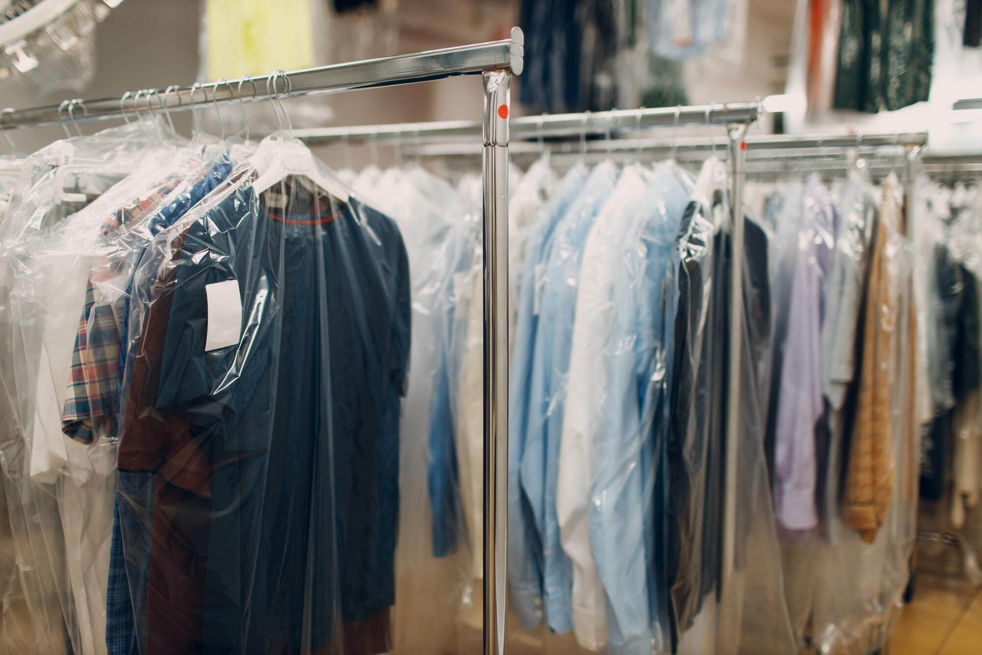 Clothes covered in clear plastic protective bags hanging on metal racks in a dry cleaning store.