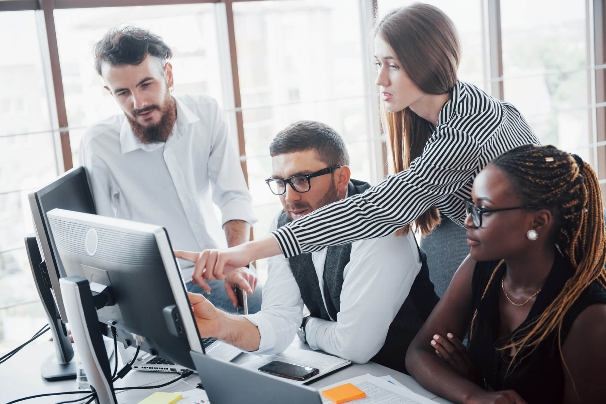 Four diverse professionals collaborating and pointing at a computer screen in a modern office.