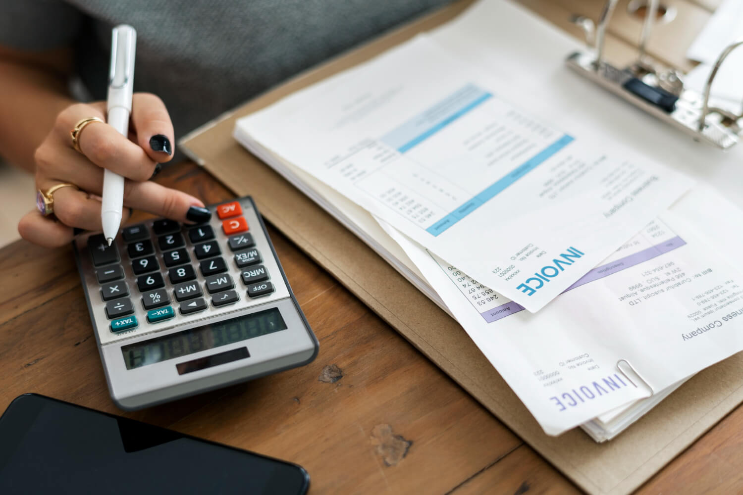 Hand with black nail polish and rings pressing buttons on a calculator next to invoices and a smartphone on a wooden table.