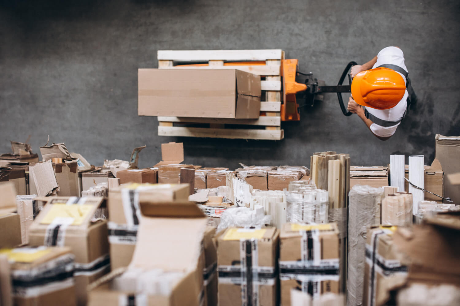 Overhead view of a worker wearing an orange hard hat operating a pallet jack to move a wooden pallet with a large cardboard box in a warehouse filled with stacked packages and wrapped goods.