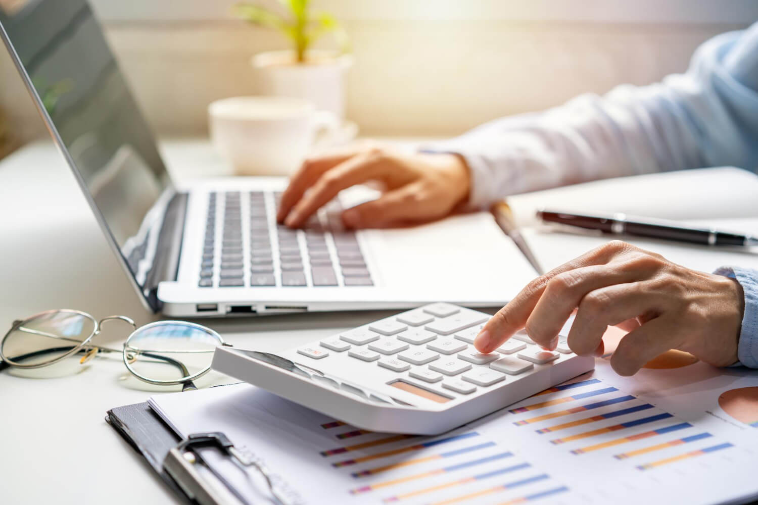 Person using a calculator and typing on a laptop with financial charts and glasses on the desk.