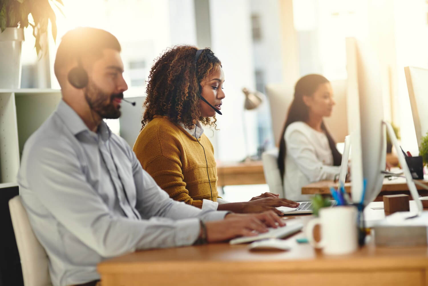Three customer service representatives wearing headsets working at computers in a bright office.