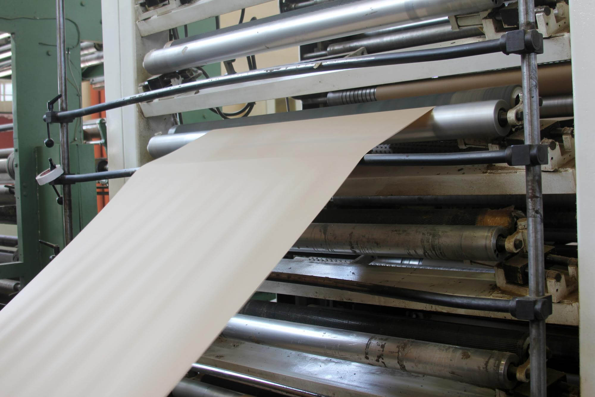 Wide sheet of beige paper being fed through rollers in an industrial machine.