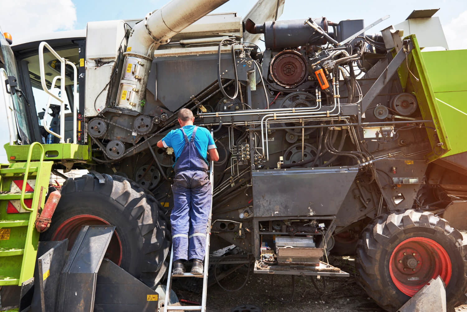 Man in blue overalls climbing a ladder to inspect or repair the inner machinery of a large agricultural harvester.