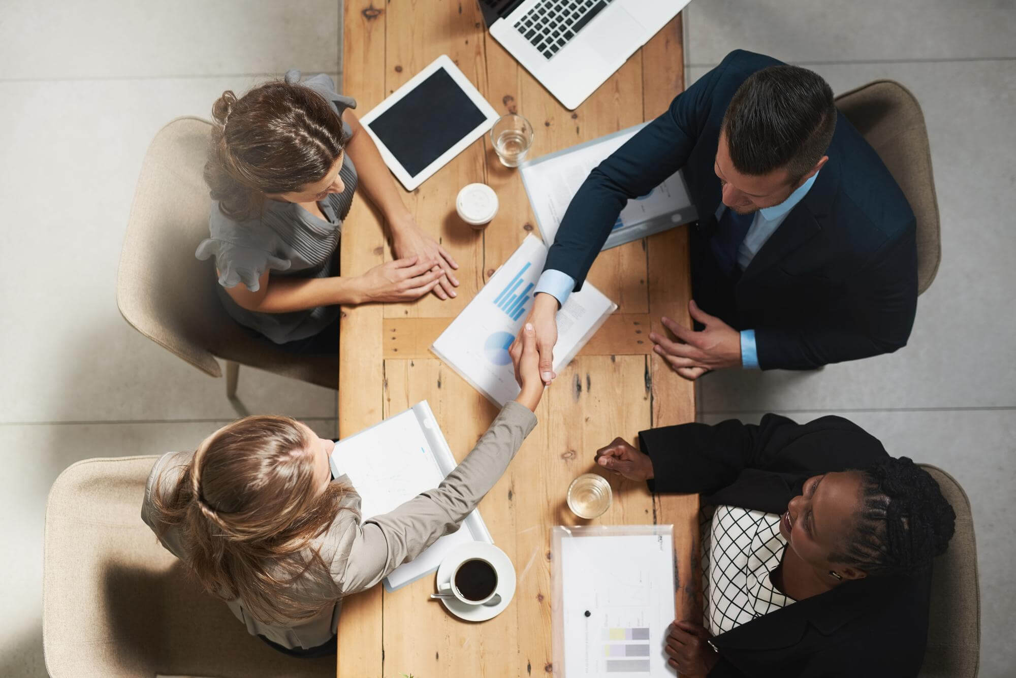 Overhead view of four businesspeople sitting around a wooden table with documents, laptops, and drinks, two shaking hands.