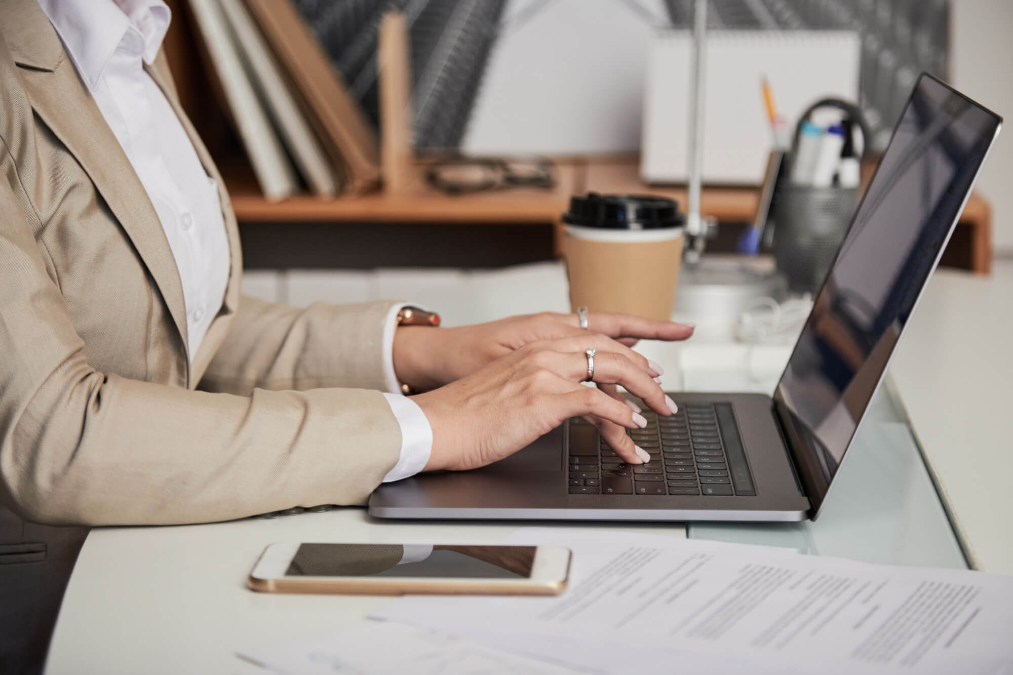 Person in beige suit typing on a laptop at a desk with a smartphone, paper documents, and a coffee cup.