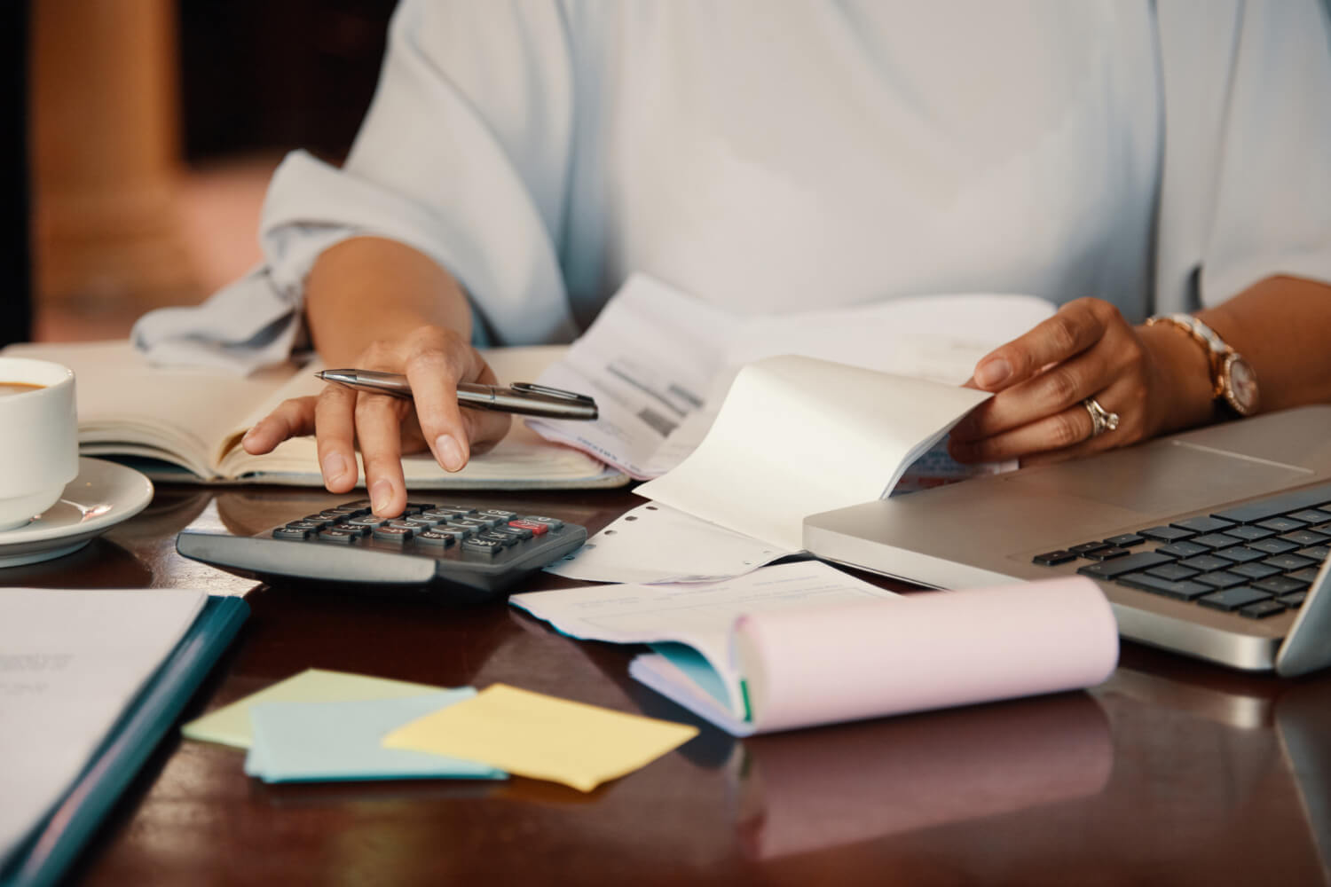 Person calculating expenses with a calculator and reviewing receipts next to a laptop on a wooden table.