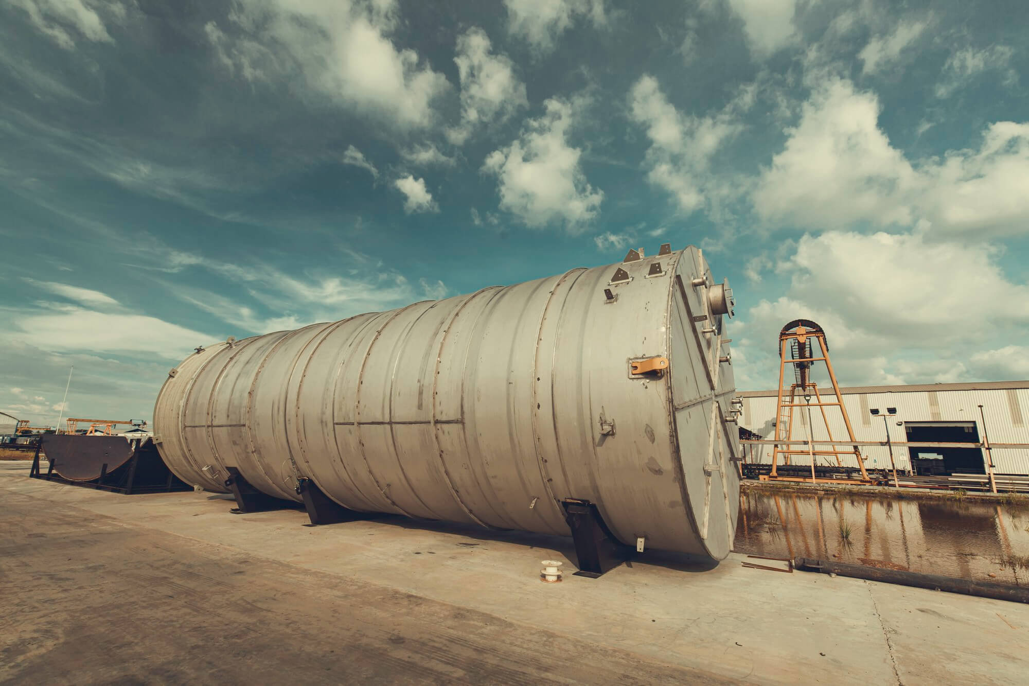 Large cylindrical industrial metal tank lying horizontally on concrete ground near a water canal with a crane structure and warehouse in the background.