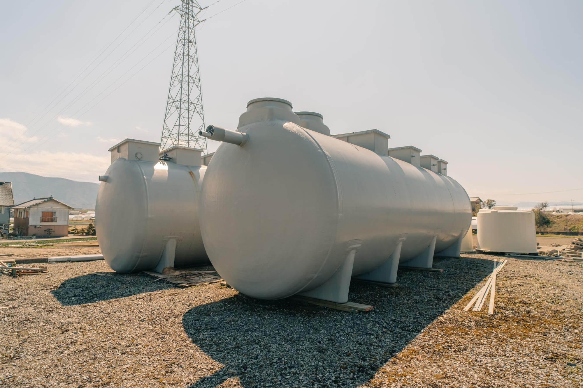 Two large industrial cylindrical storage tanks placed outdoors on gravel ground with small buildings and electrical tower in the background.