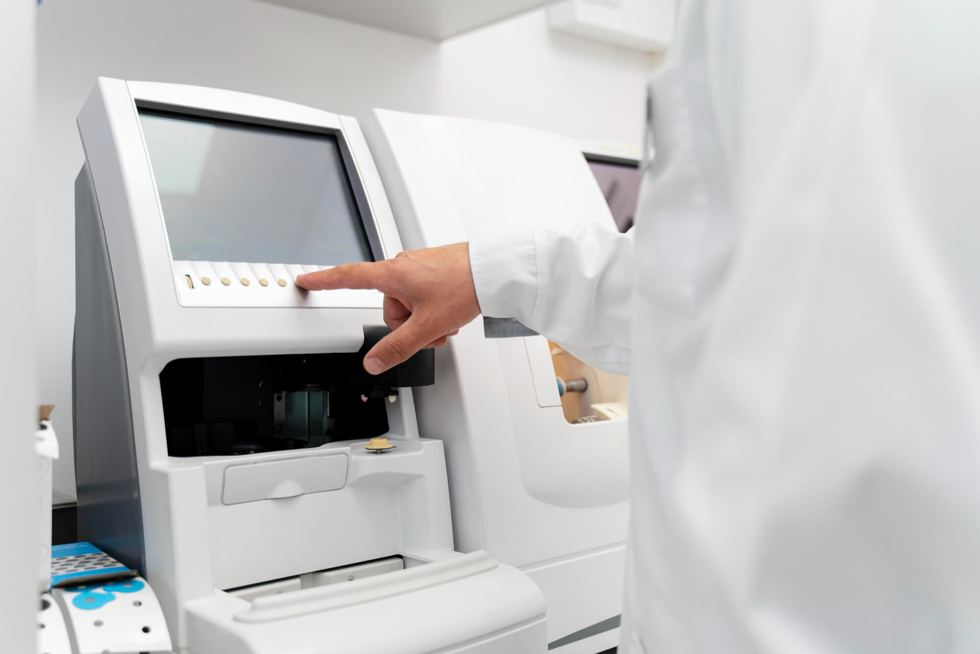 Person in a white lab coat pressing buttons on a laboratory instrument with a screen.