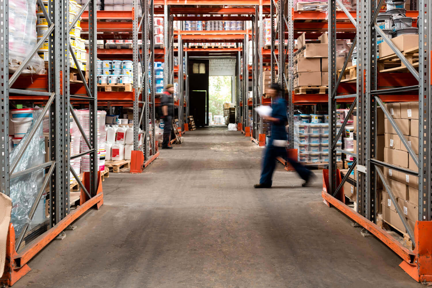 Warehouse aisle with shelves stocked with boxes and containers, and two blurred workers in motion.