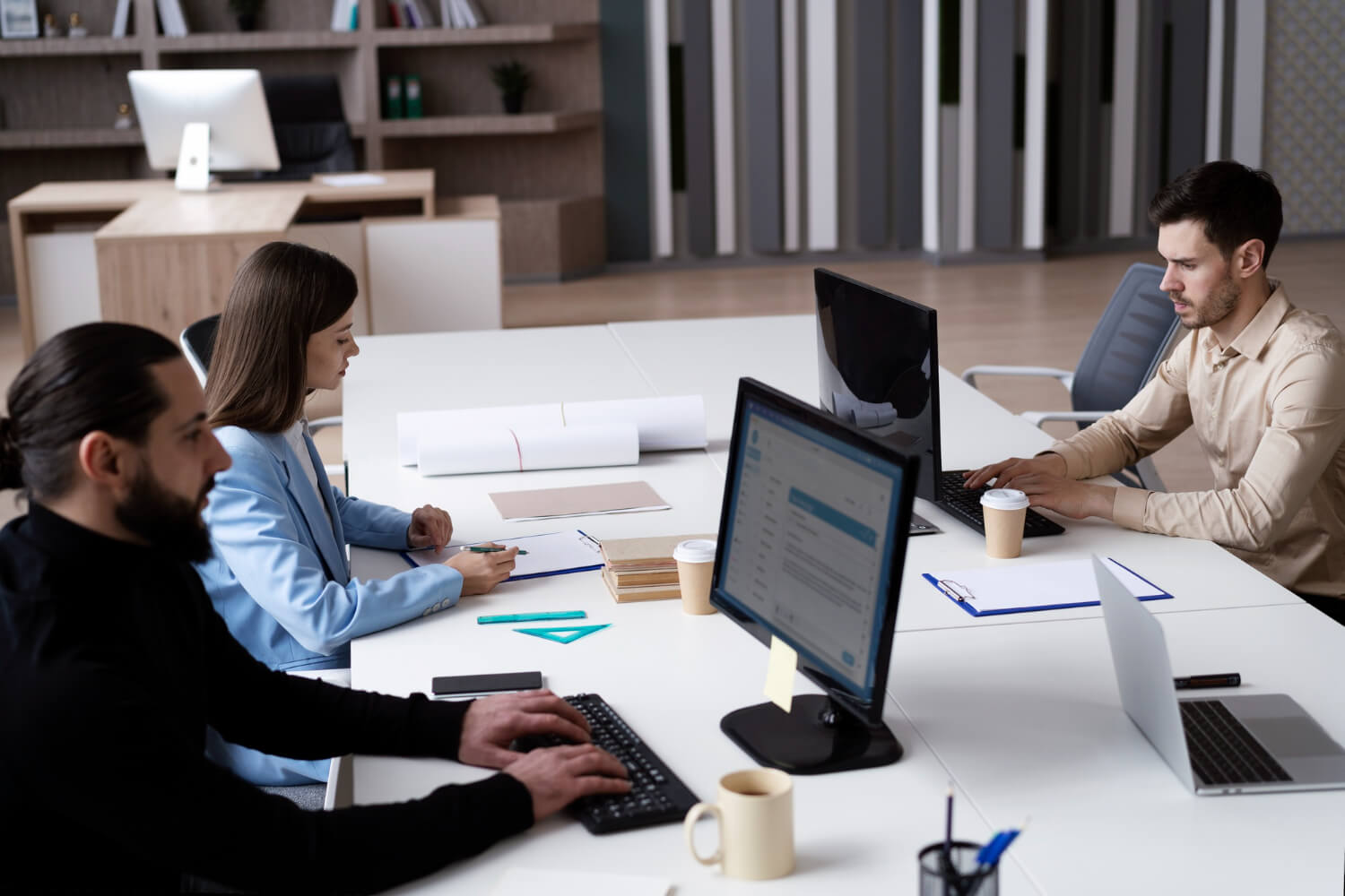 Three coworkers focused on work at a large white desk with computers, notebooks, and coffee cups in a modern office.