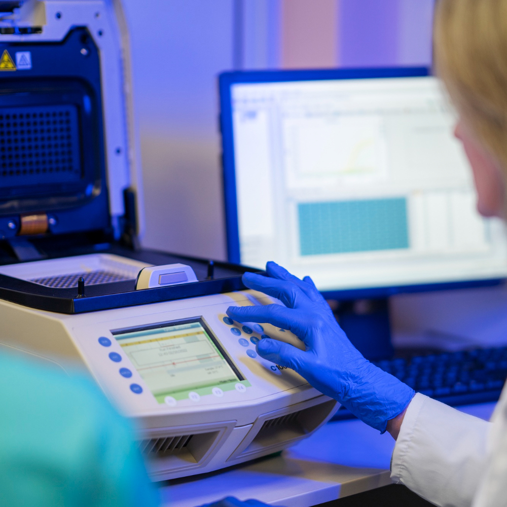 Scientist wearing blue gloves operating a laboratory thermal cycler machine with a computer screen displaying data in the background.