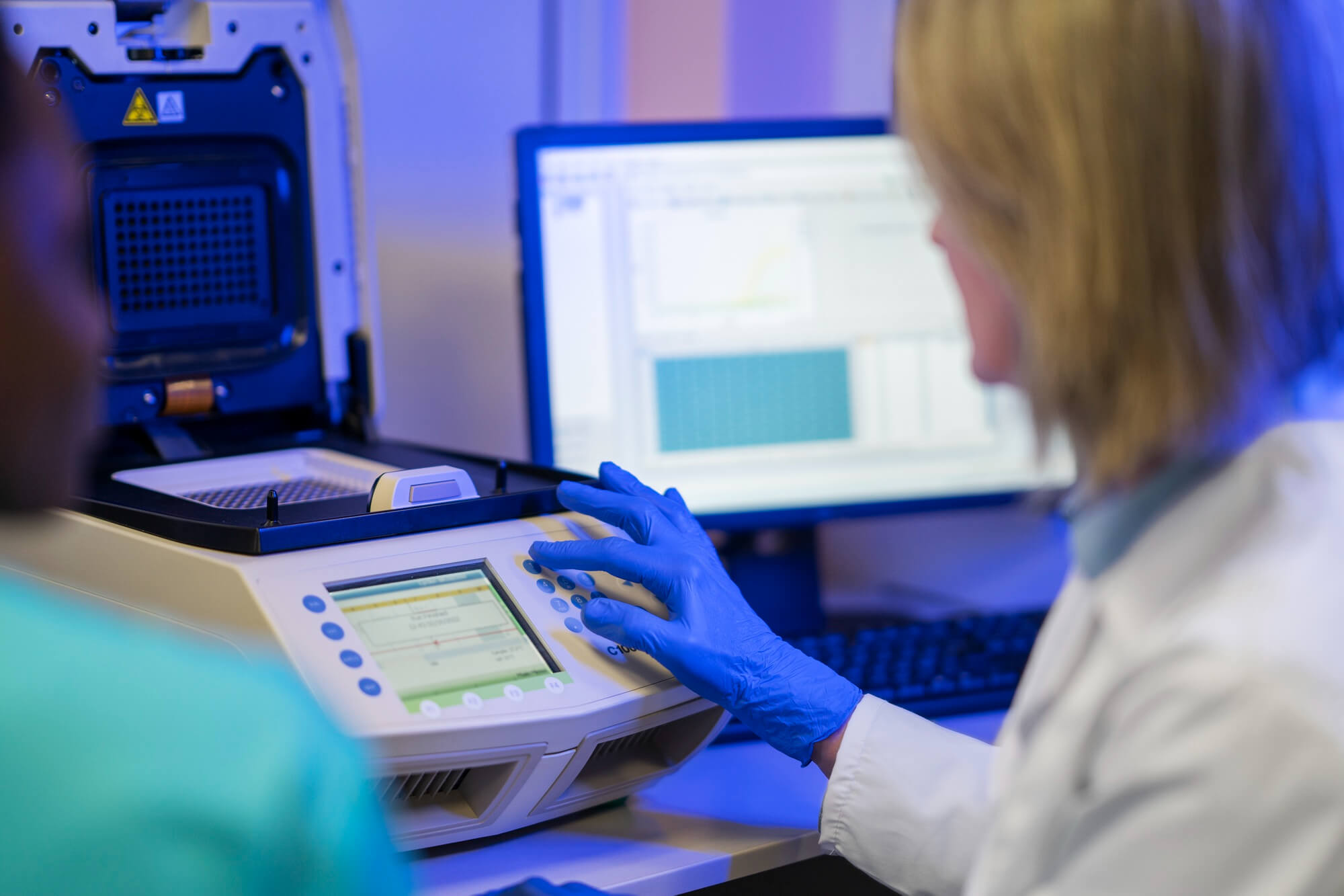Scientist wearing blue gloves operating a laboratory thermal cycler machine with a computer monitor displaying data in the background.