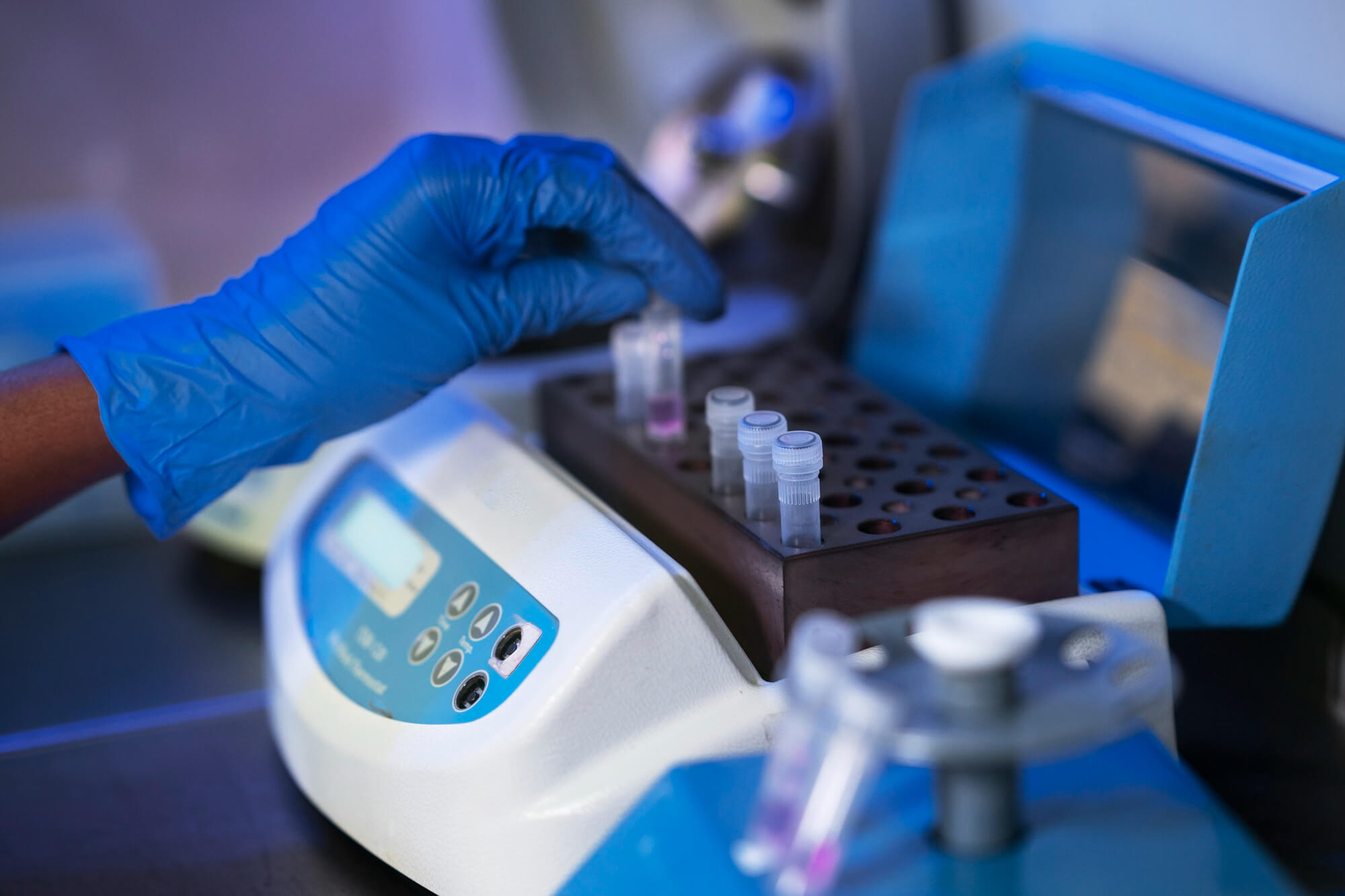Gloved hand placing a small vial with pink liquid into a lab centrifuge machine's vial holder.