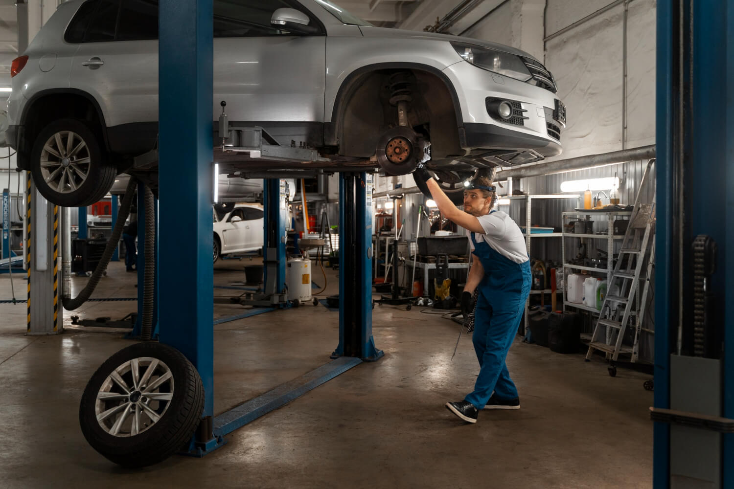 Mechanic inspecting the underside of a silver car lifted on a hydraulic lift in a garage.