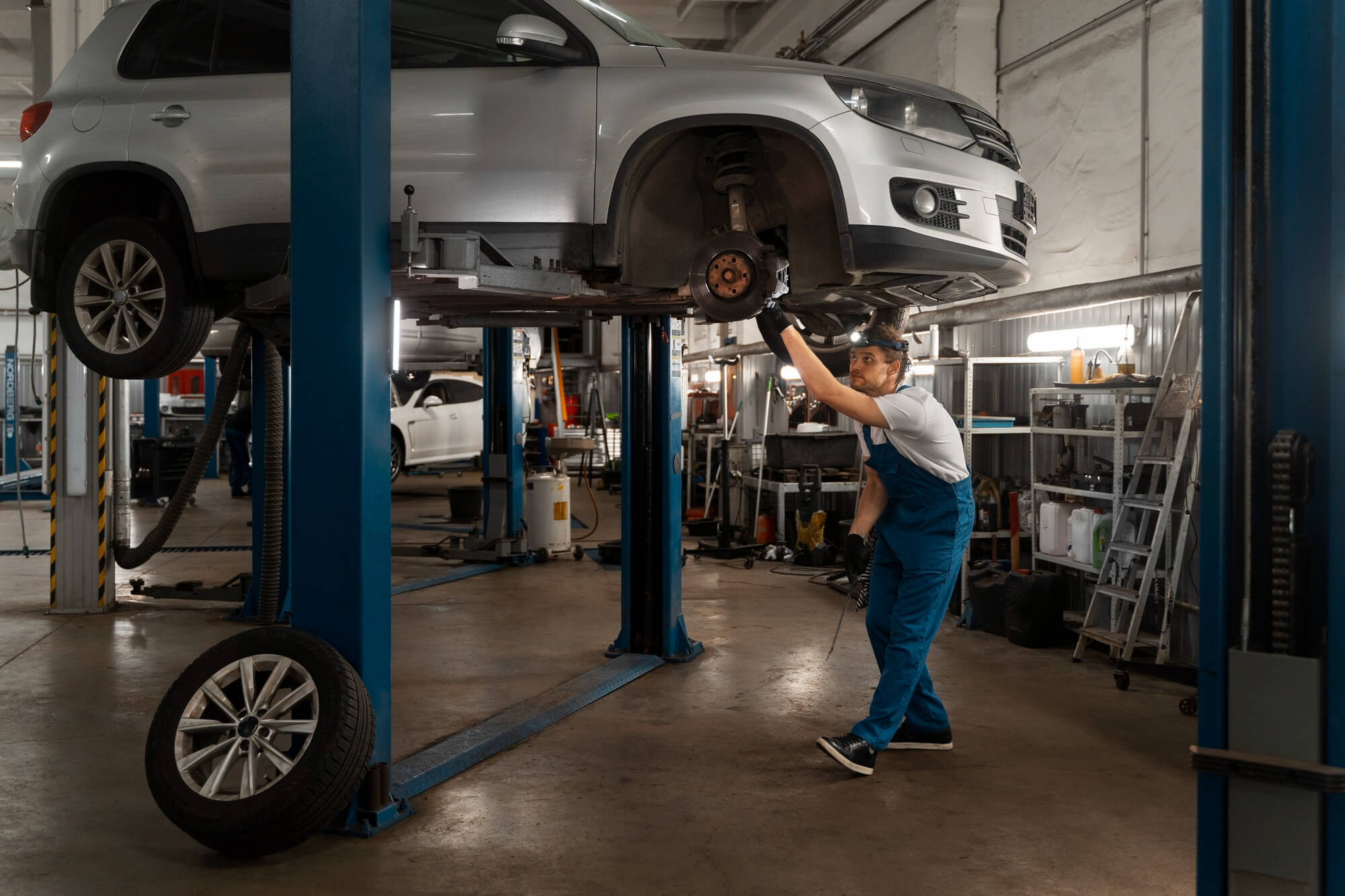 Mechanic in blue overalls repairing a silver car lifted on a hydraulic lift inside a garage.