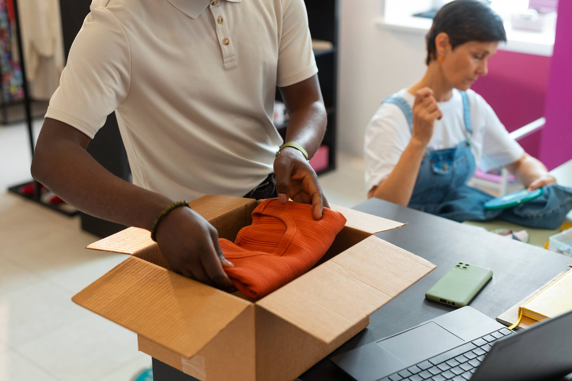 Person packing an orange folded sweater into a cardboard box with a laptop and phone on the table, another person in background handling denim clothing.