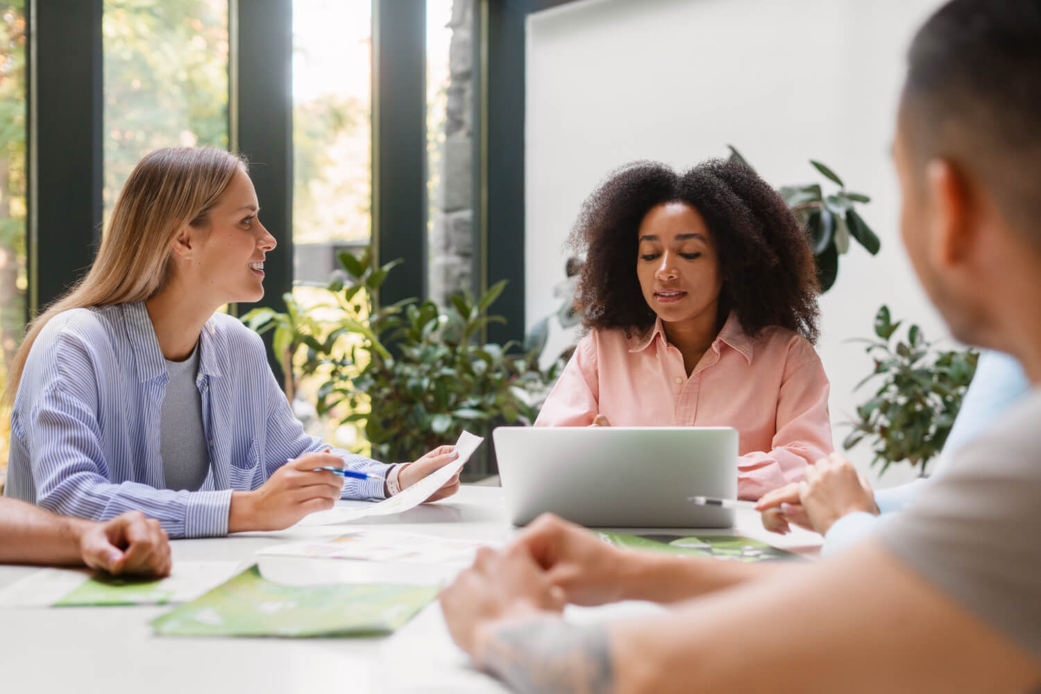 Four diverse colleagues engaged in a meeting around a table with laptops and documents in a bright office with plants.