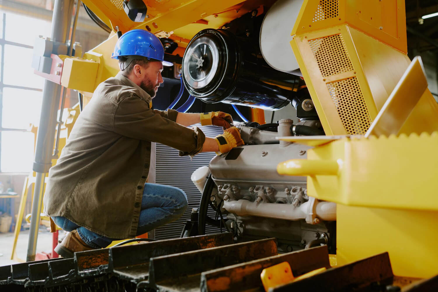 Male mechanic wearing a blue hard hat and gloves working on the engine of heavy yellow machinery inside a workshop.