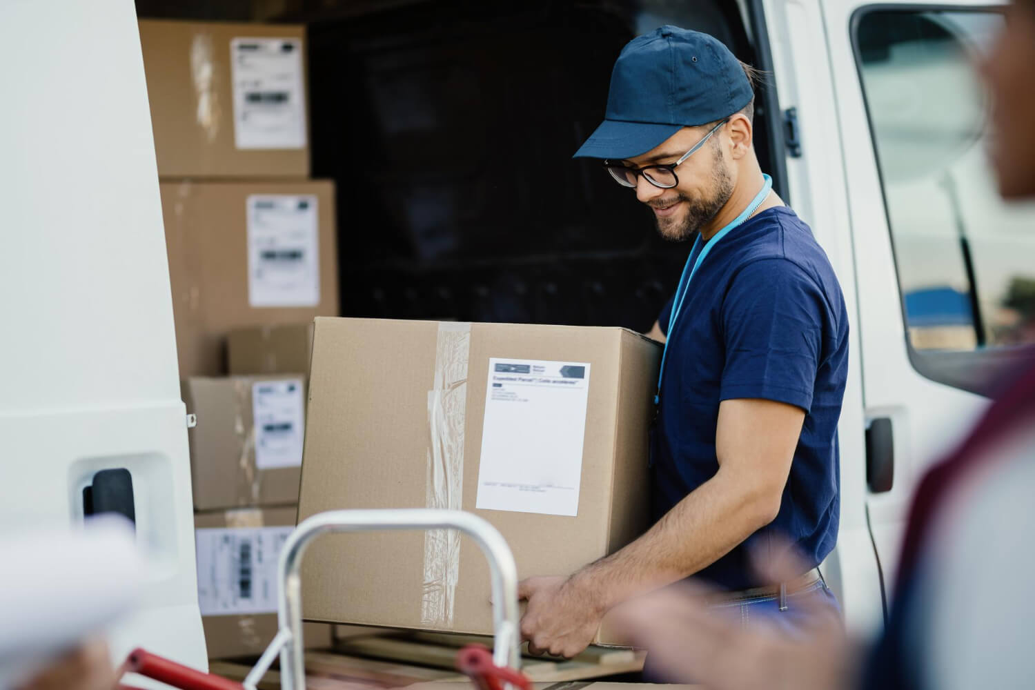 Delivery worker wearing a blue cap and glasses carrying a large cardboard box near a delivery van.