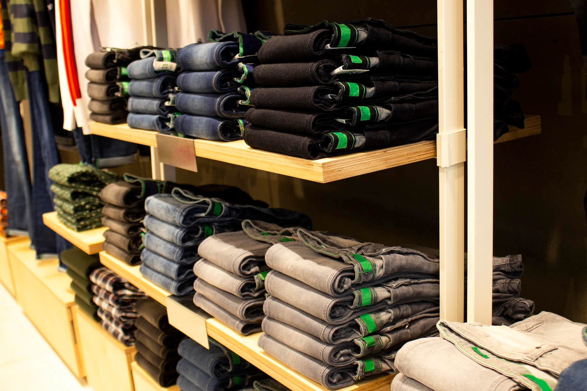 Stacks of folded jeans in various colors on wooden shelves in a clothing store.