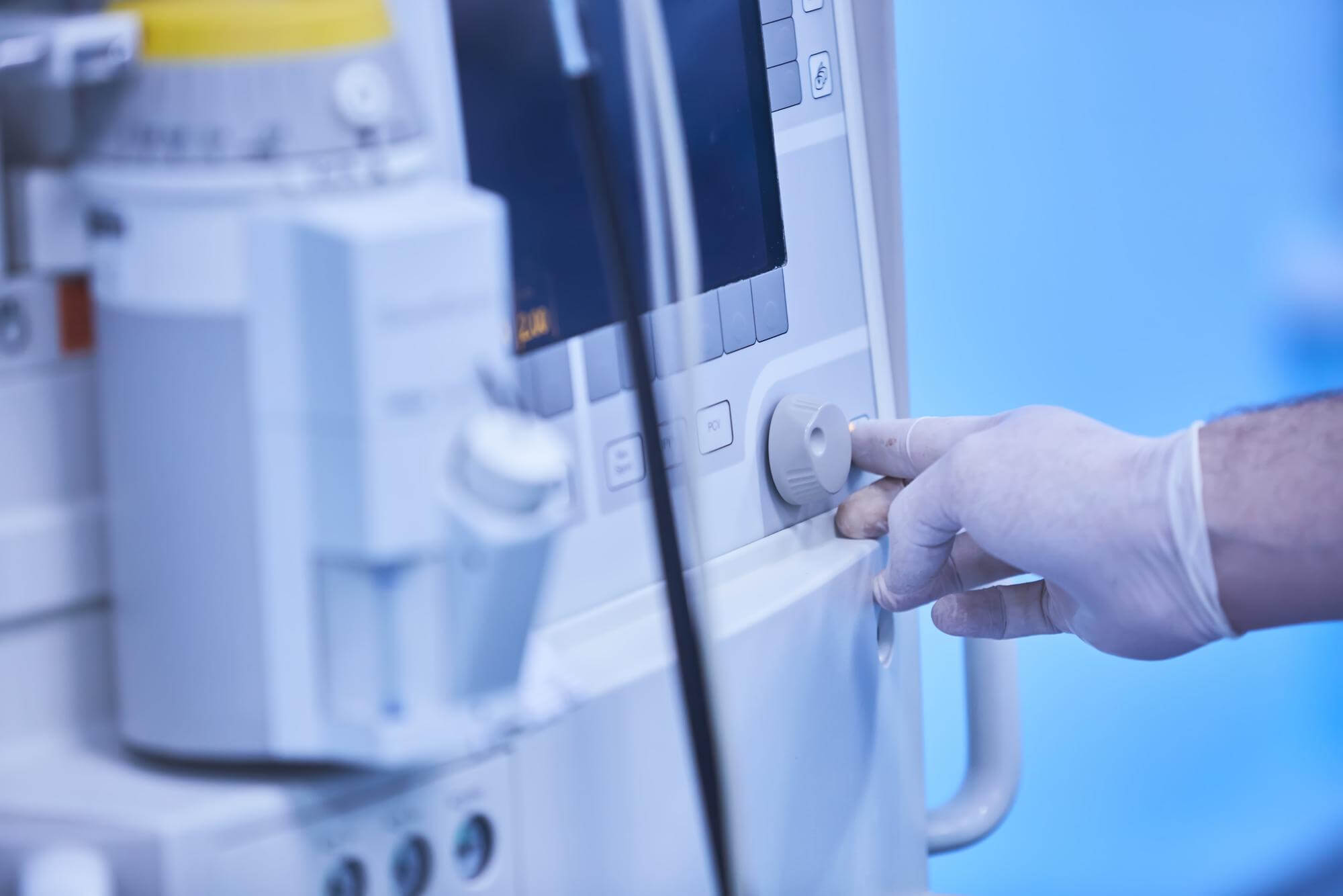 Hand in a medical glove adjusting a knob on hospital medical equipment with a screen.