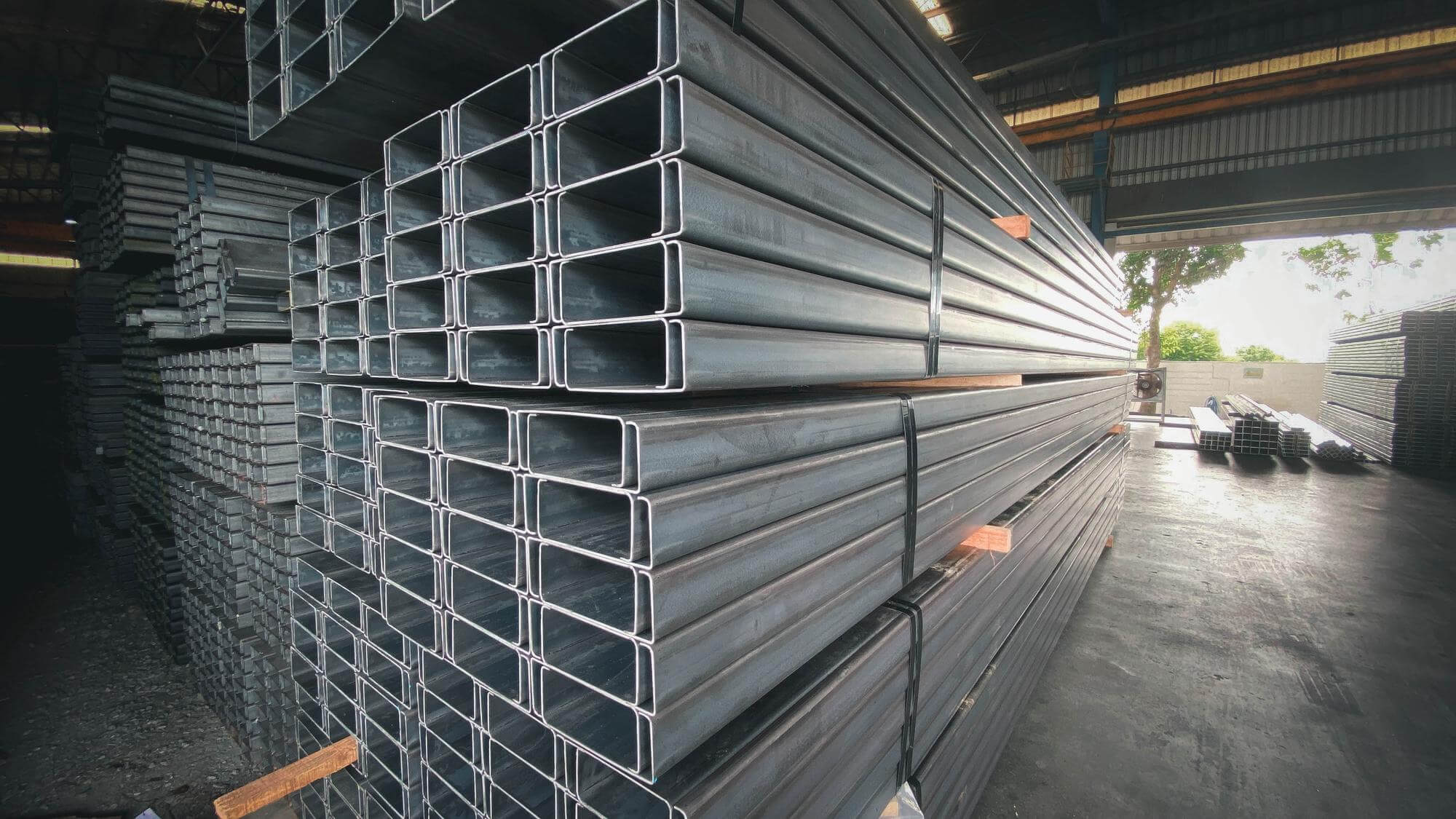 Stacks of large rectangular steel pipes stored inside a warehouse with an open side showing outdoor trees.