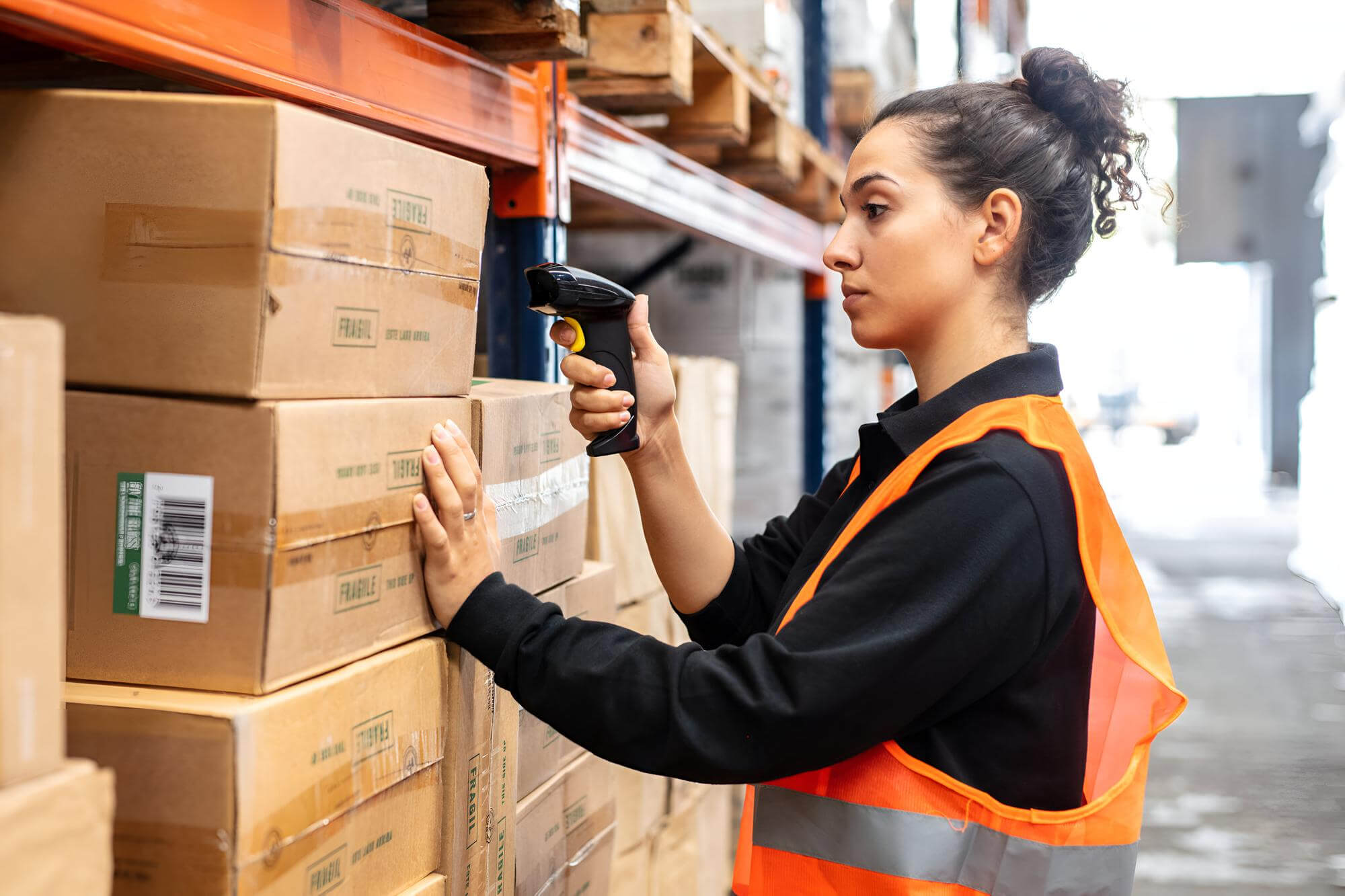Woman wearing an orange safety vest using a barcode scanner to scan boxes stacked on warehouse shelves.