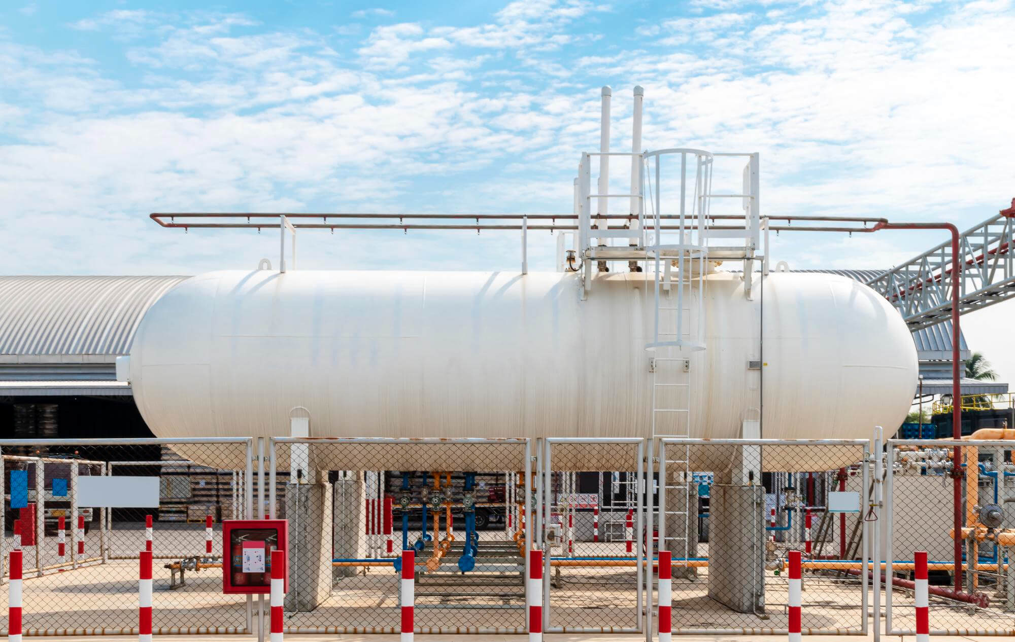 Large white industrial storage tank elevated on concrete supports with metal fencing and pipes underneath.