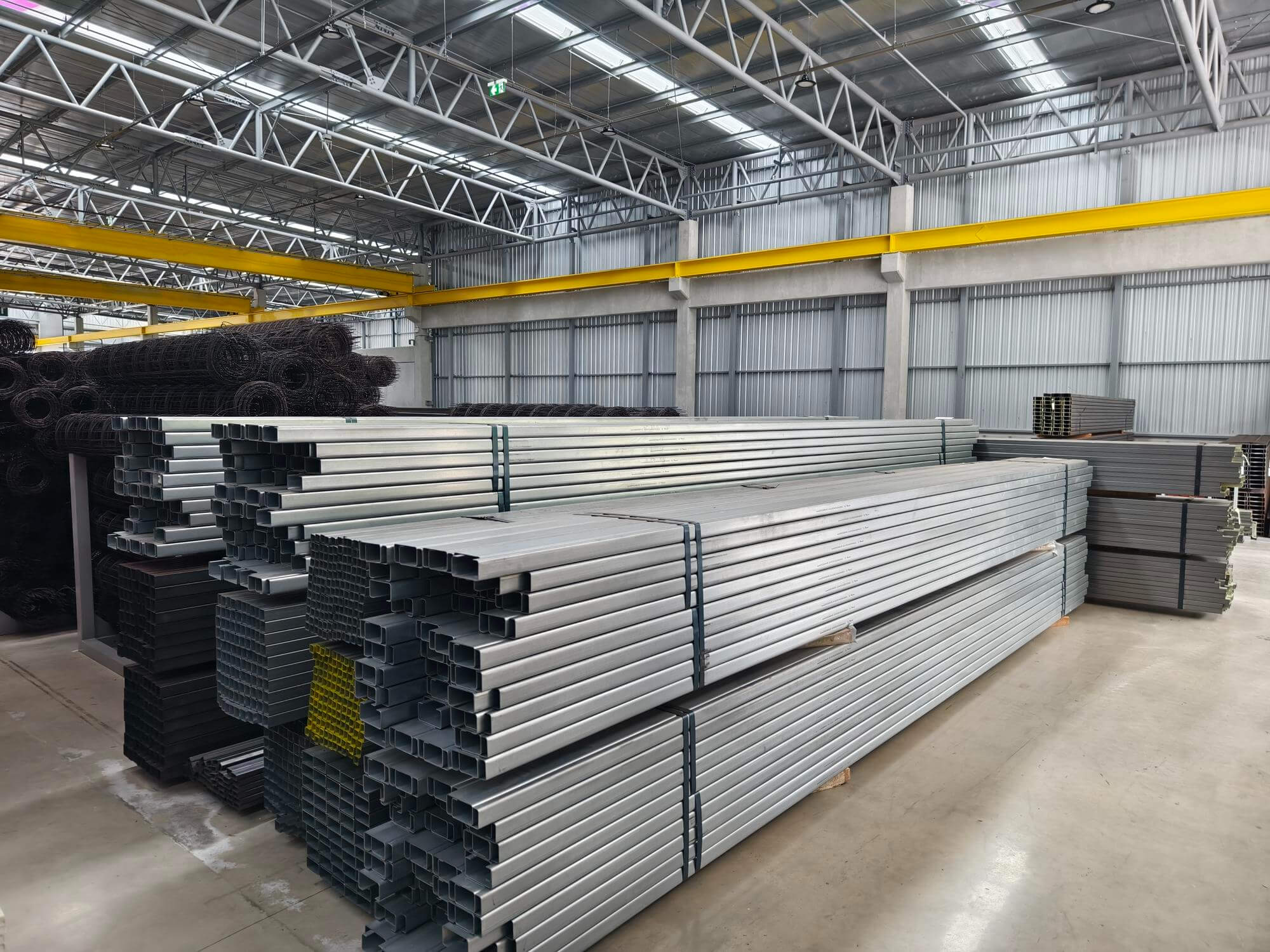 Stacked rectangular steel pipes and wire mesh rolls stored inside a spacious metal warehouse.