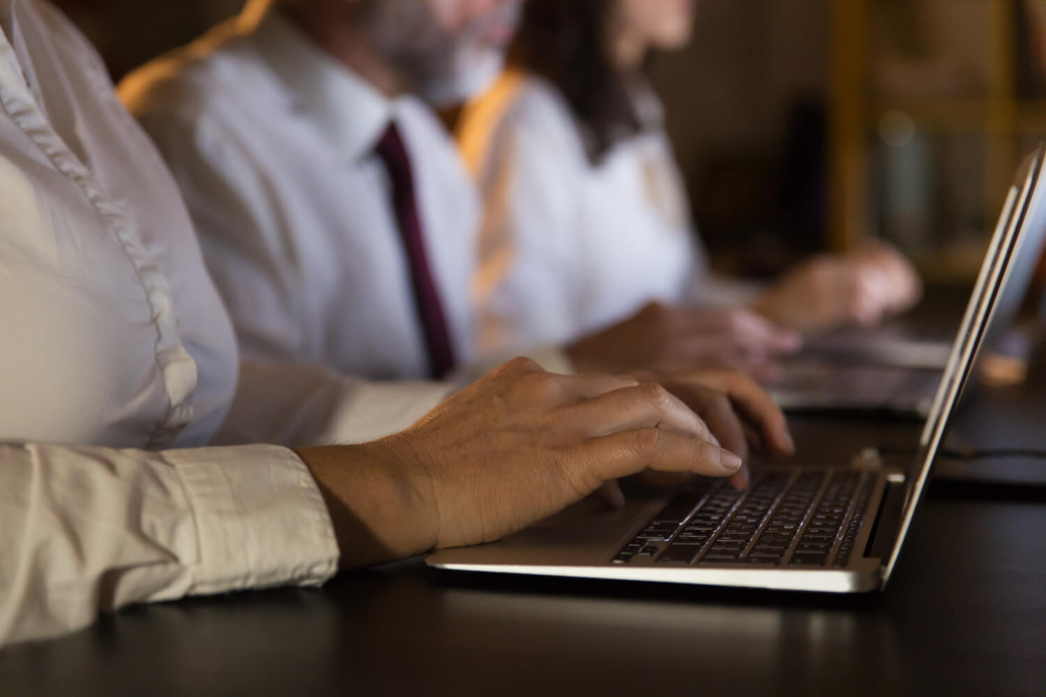 Close-up of people in business attire typing on laptop keyboards in a dimly lit setting.