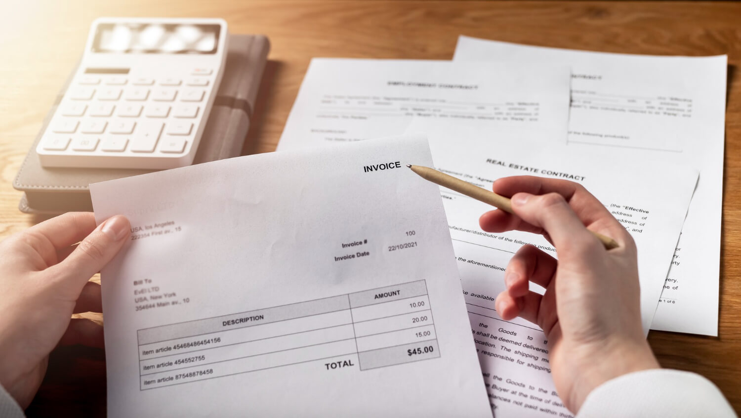 Person holding an invoice with item descriptions and amounts totaling $45, with a calculator and contract papers on a wooden desk.