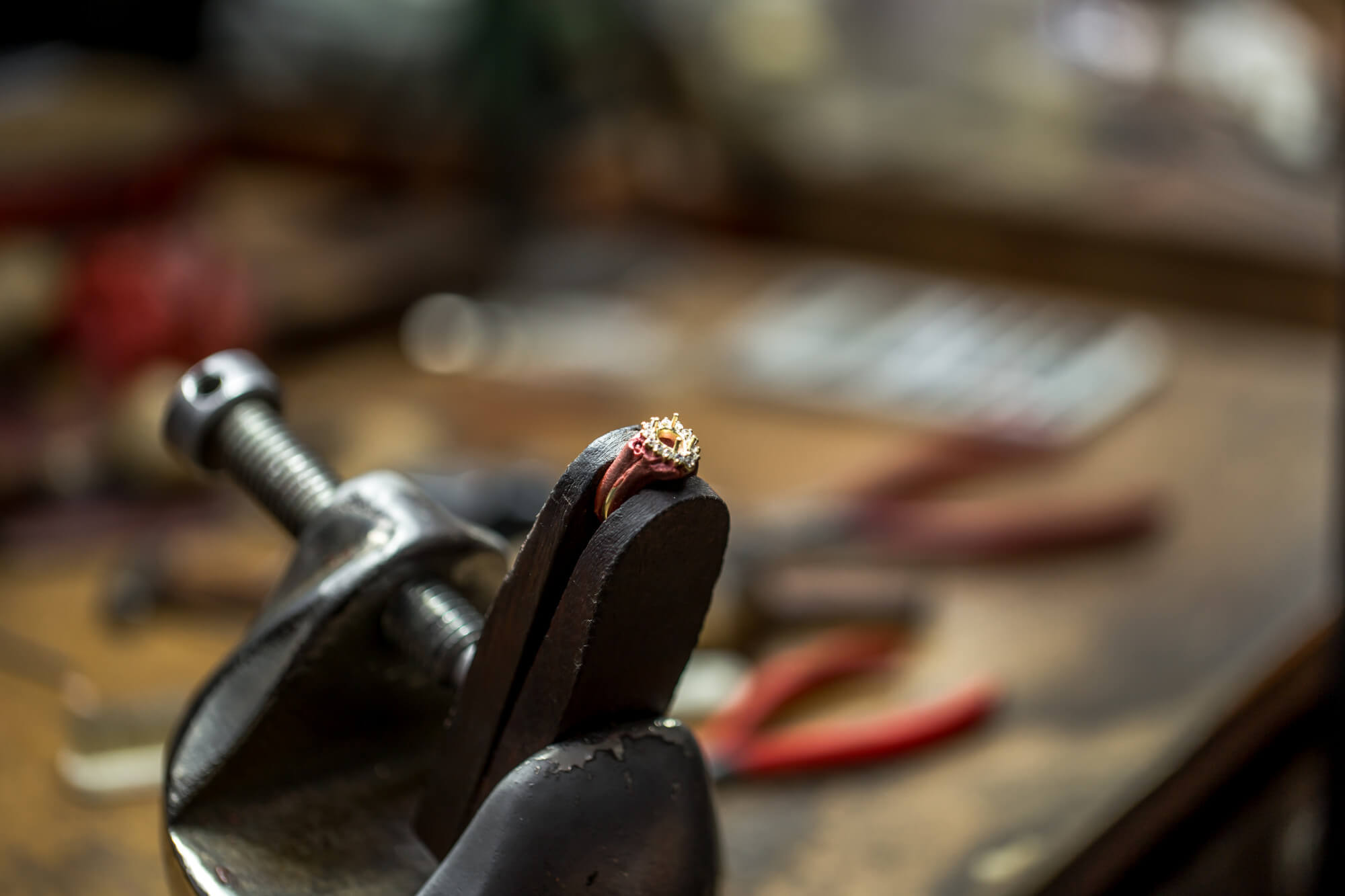 A delicate ring with a gemstone held in a metal vise on a jeweler's workbench with blurred tools in the background.