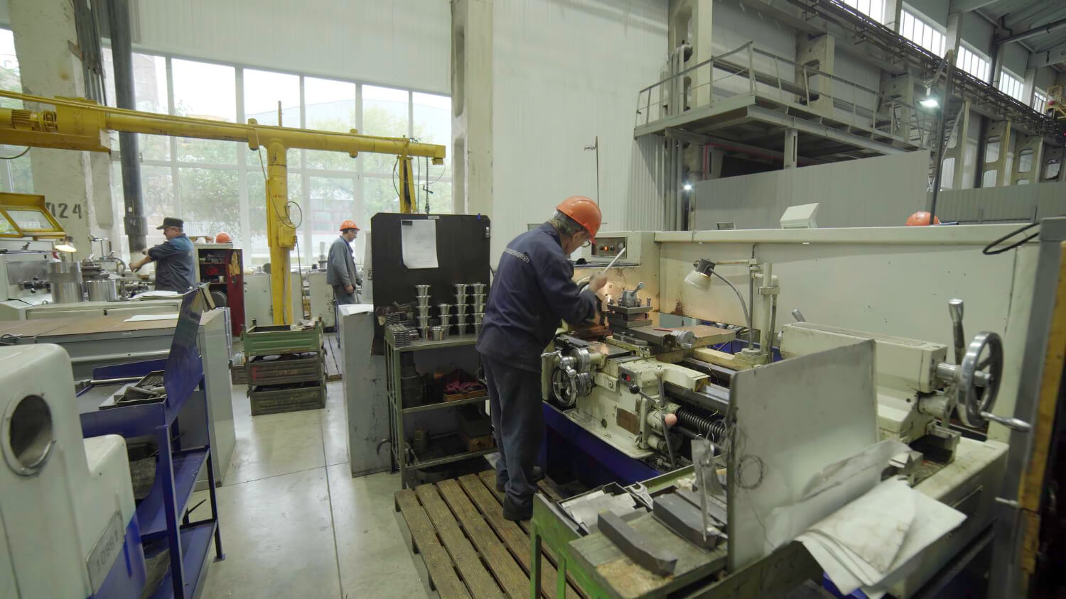 Workers wearing orange helmets operating machinery inside a bright industrial workshop.