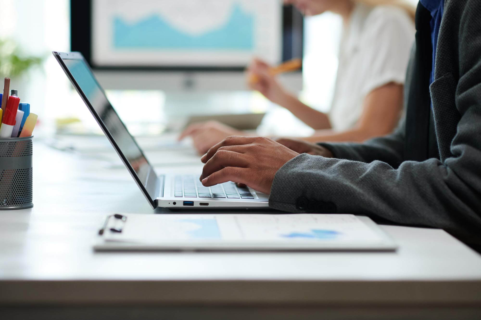 Person typing on a laptop at a desk with office supplies and a document, another person blurred in the background working.